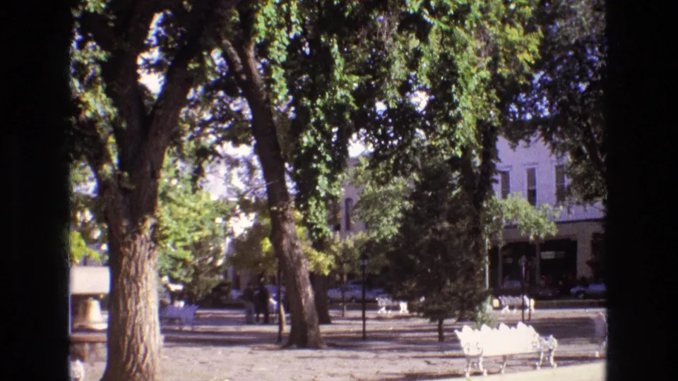 1973 New Mexico home-movie frame showing a quiet plaza under large leafy trees, with white benches, parked cars, and storefront-like buildings in the background.