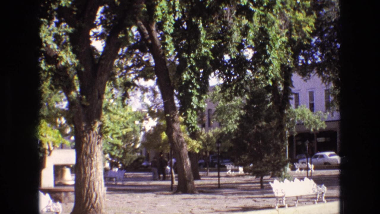 1973 New Mexico footage frame of a small-town plaza with mature trees, empty white benches, parked cars, and low commercial buildings beyond the street.