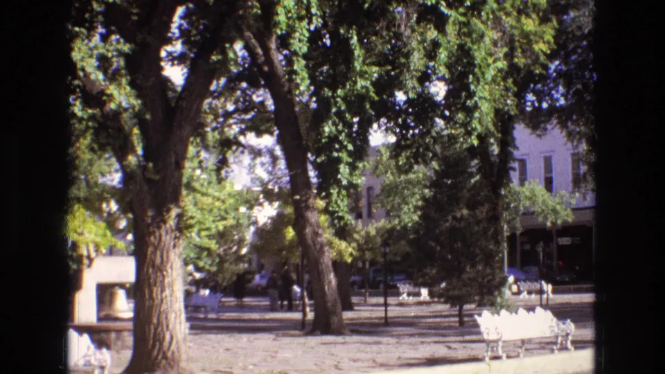 1973 New Mexico home-movie frame with tall shade trees, white plaza benches, distant pedestrians, parked cars, and background buildings along the street.