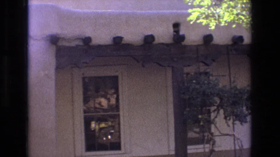 1973 New Mexico home-movie frame shifting toward a nearby building exterior with a window, projecting roof beams, foliage, and a partial edge of the plaza scene.