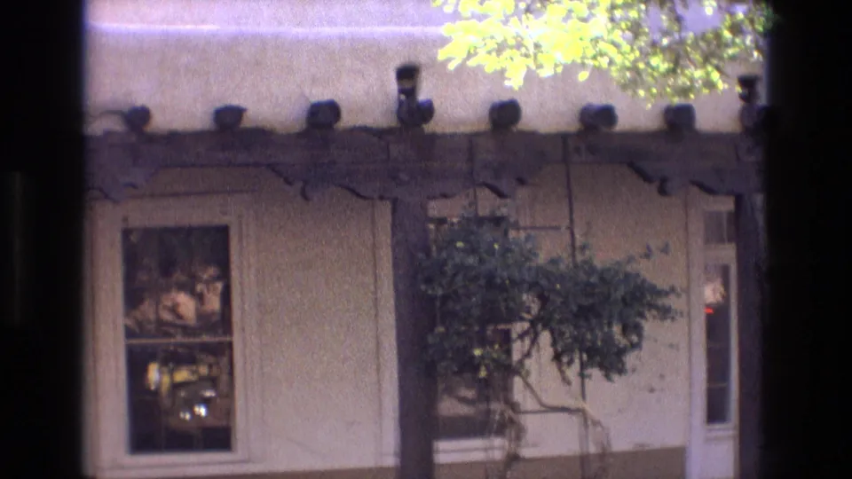 1973 New Mexico home-movie frame showing an adobe-style exterior with a wooden porch beam, a window, a glass-paneled door, and leafy courtyard planting.