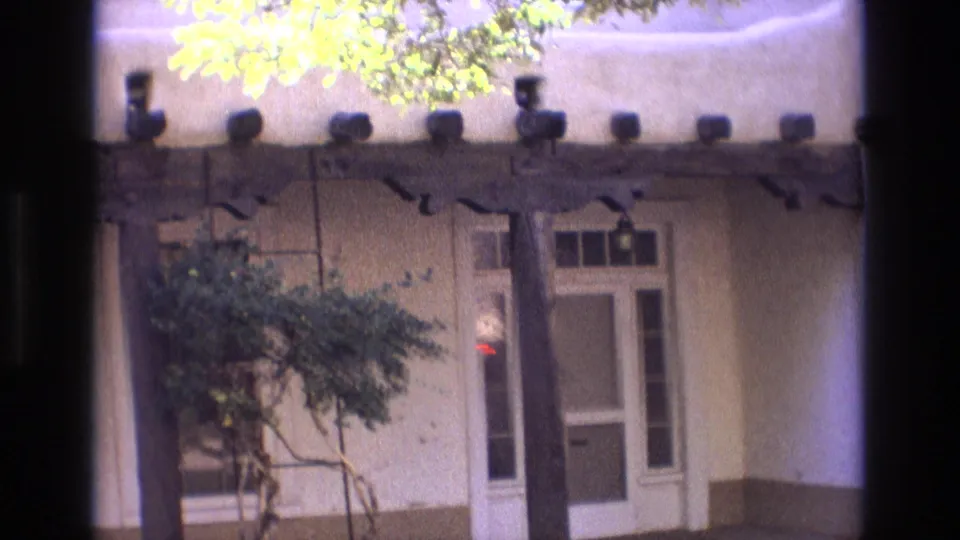 1973 New Mexico home-movie frame showing the entrance area from a slightly shifted angle, with a small tree, porch posts, window, and glass-paneled door.