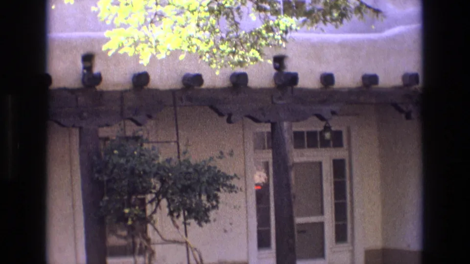 1973 New Mexico exterior frame showing an adobe-style house frontage with a heavy wooden beam, courtyard tree, window, white door, and sunlit leaves overhead.