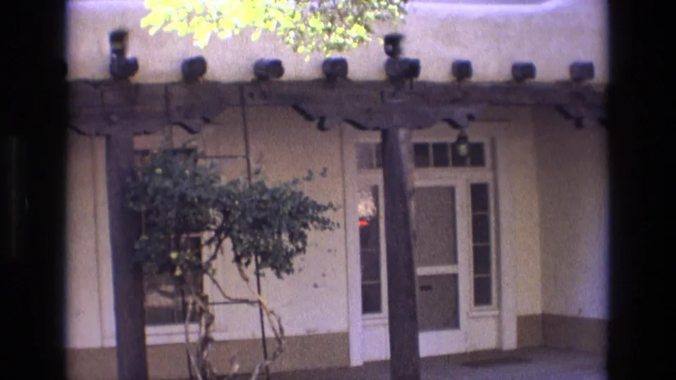 1973 New Mexico home-movie frame showing an adobe-style building facade with vigas, a white entry door, narrow glass panels, and a small tree near the wall.