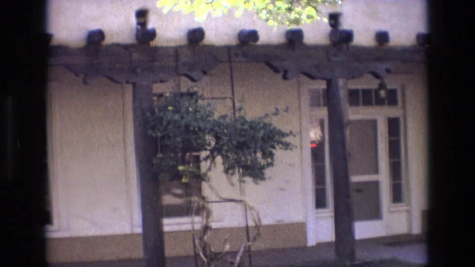 1973 New Mexico frame of an adobe-style exterior with exposed wooden beams, a pale door, side windows, and a tree partly covering the wall.