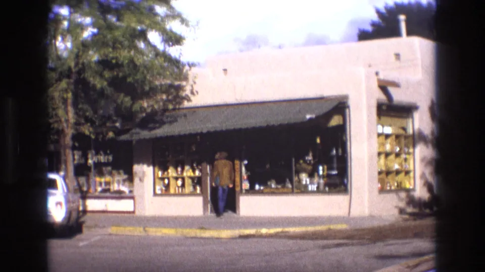 1973 New Mexico street view showing a low stucco storefront with large display windows, a dark awning, tree shade, and a curb at the corner.