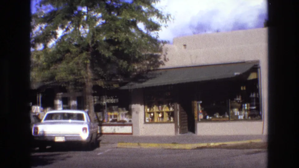 1973 New Mexico street frame with a white classic American car parked beside a shaded storefront with display windows, a dark awning, and trees overhead.