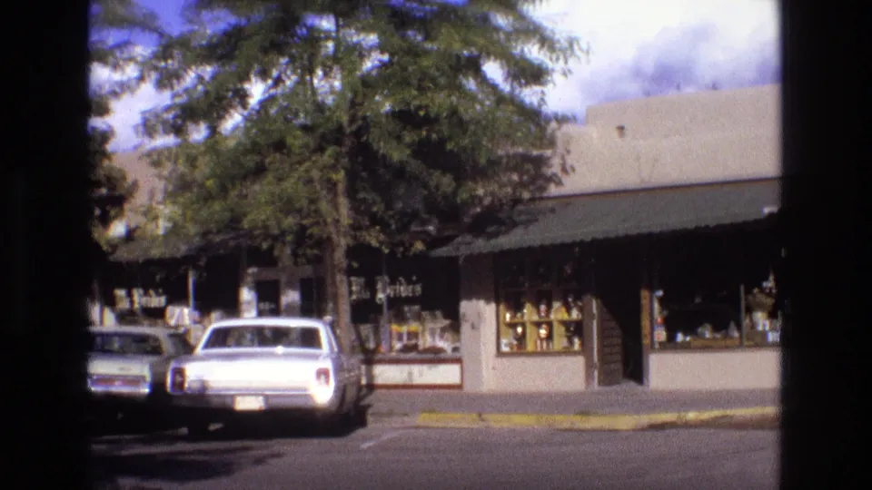 1973 New Mexico 8mm frame showing a sunlit downtown storefront, leafy trees, parked cars, and a white car along the curb.