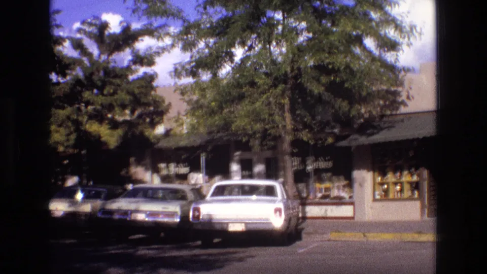 1973 New Mexico street view with storefront windows, mature trees, a white parked car, and other automobiles along the curb.