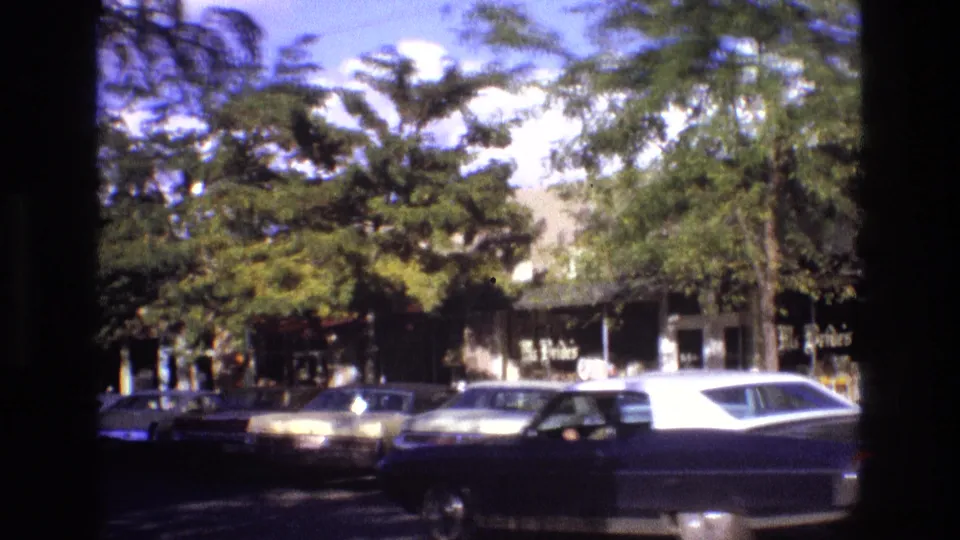 1973 New Mexico 8mm frame with cars moving past a shaded downtown shopping block, parked vehicles, trees, and partially visible signs.