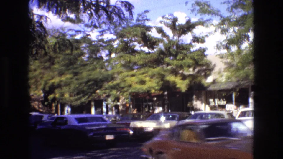 1973 New Mexico downtown street frame showing moving traffic, parked cars, leafy trees, shop windows, and partially readable storefront signage.