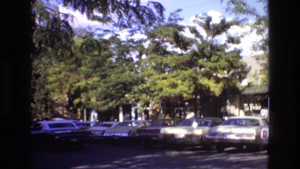 1973 New Mexico 8mm frame showing classic cars parked along a tree-lined street, with low buildings partly visible beyond the vehicles.