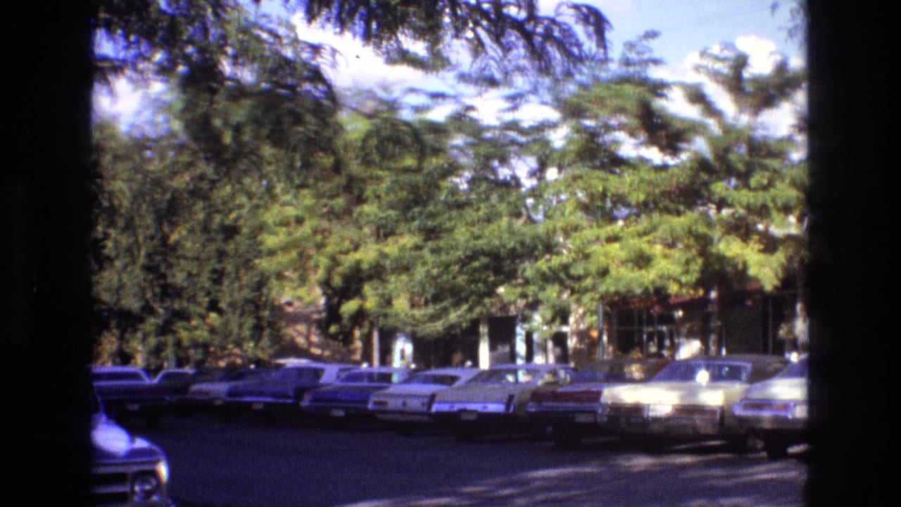 1973 New Mexico street view with period cars parked along both sides, partially obscured by trees and dark frame-edge shadows from the 8mm scan.