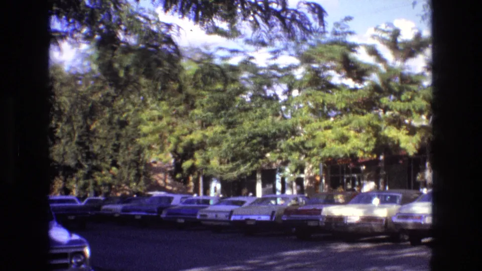 1973 New Mexico street view with period cars parked along both sides, partially obscured by trees and dark frame-edge shadows from the 8mm scan.