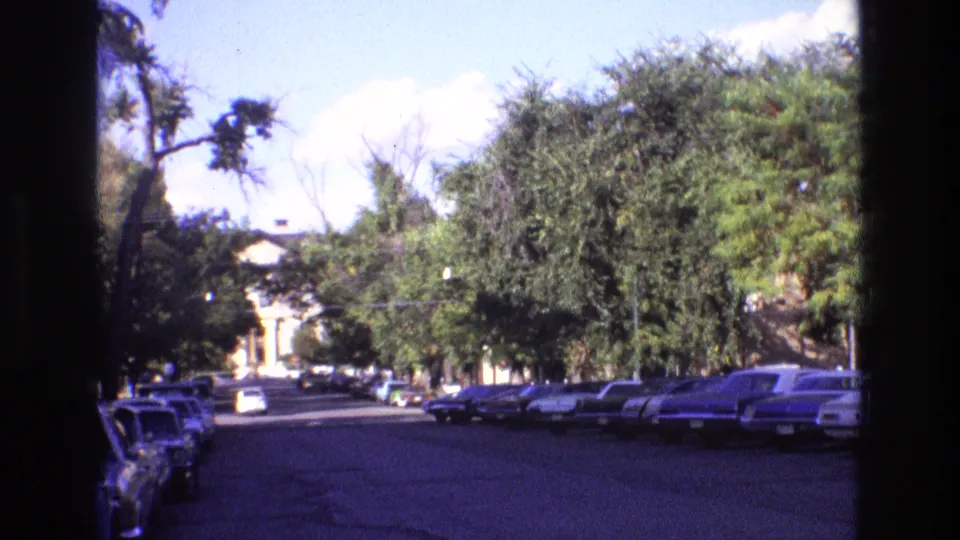 1973 New Mexico 8mm view down a sunlit street lined with parked cars, mature trees, and a distant light-colored building or dome-like form.