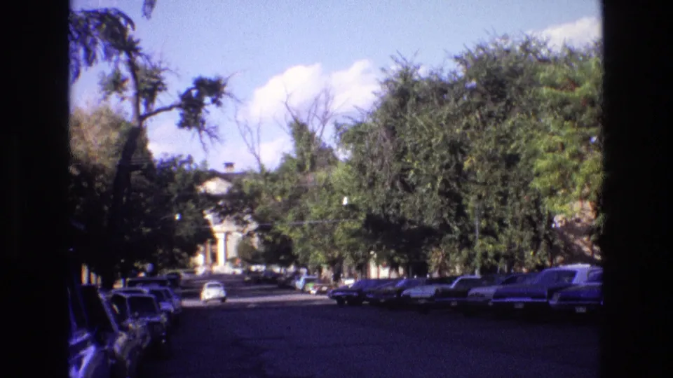 1973 New Mexico home-movie frame looking along a quiet street, with parked period cars, heavy tree cover, and a distant pale building or dome-like feature.