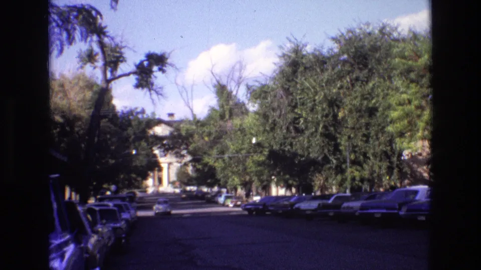 1973 New Mexico 8mm frame showing a wide street bordered by parked cars, mature trees, blue sky, and a vehicle approaching in the distance.
