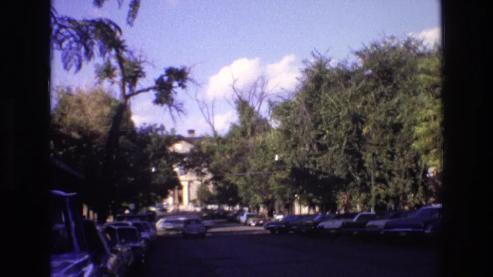 1973 New Mexico home-movie frame of a broad street where an approaching car passes between parked vehicles and shaded trees.