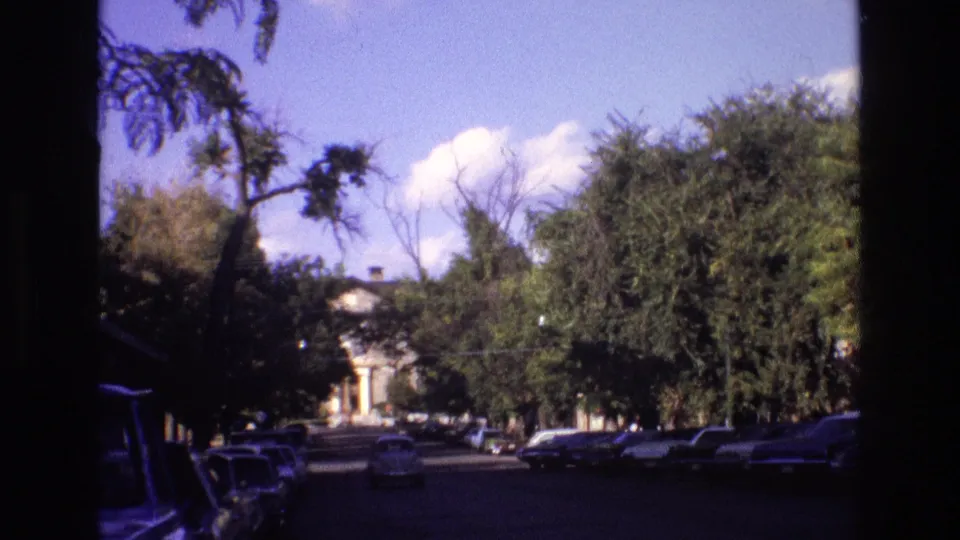 1973 New Mexico 8mm street scene with dark edge framing, parked cars, leafy trees, and a vehicle approaching under a bright sky.