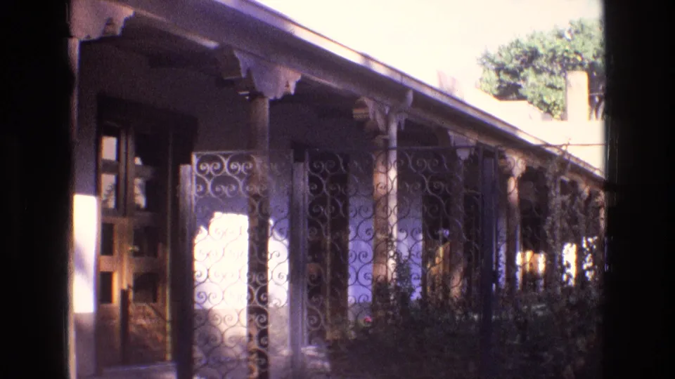 1973 New Mexico home-movie frame showing a sunlit adobe-style porch viewed past decorative spiral iron gate panels, with wooden columns, shrubs, and a doorway at left.