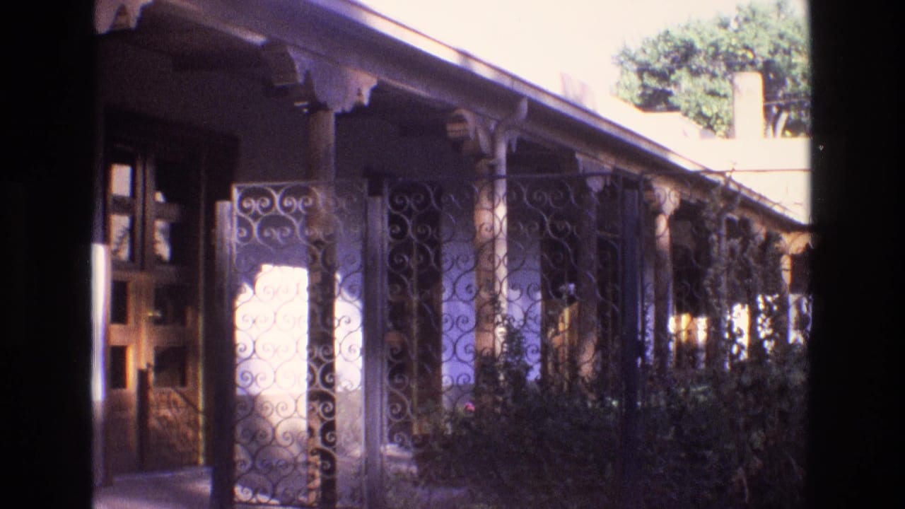 Long porch with decorative iron gate, columns, stucco wall, shrubs, and trees.