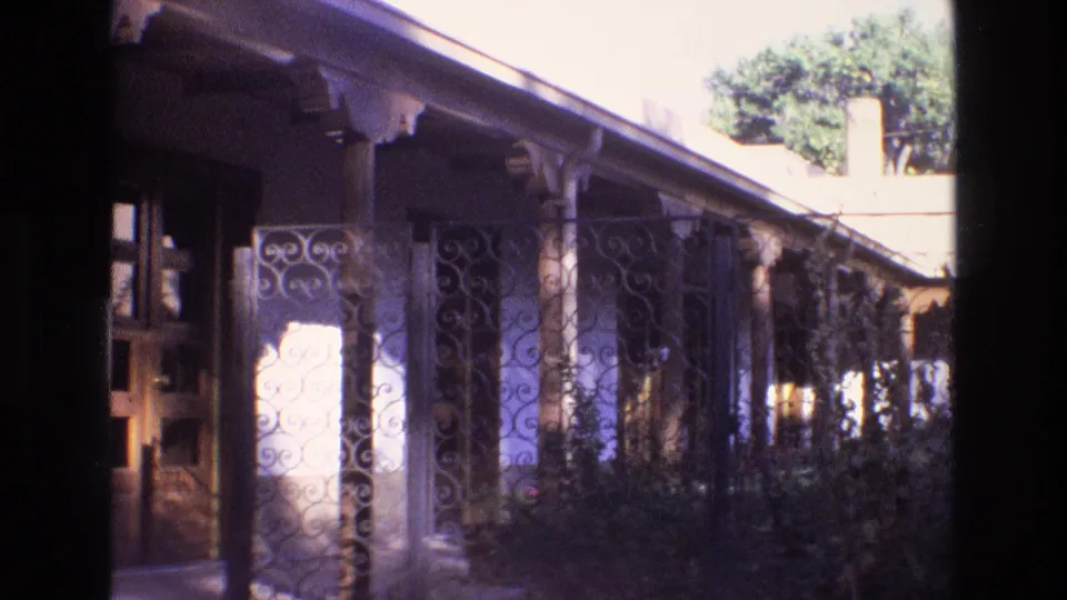 1973 New Mexico 8mm frame showing a residential porch with a doorway at left, spiral ironwork in the center, wooden columns, foliage, and bright sun across the wall.
