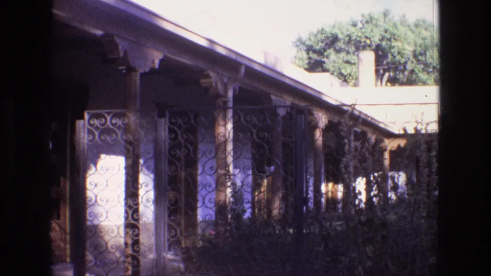 1973 New Mexico home-movie frame showing the shaded length of an adobe-style porch with wrought iron gate panels, columns, shrubs, roof eave, and trees in the background.