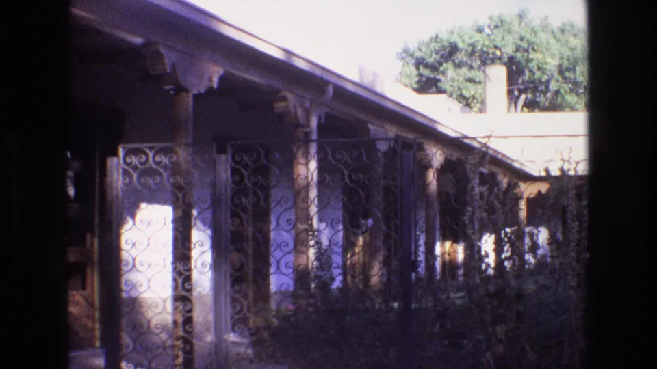1973 New Mexico 8mm frame showing a shaded resort walkway with wrought iron scrollwork, stone columns, courtyard plants, and sunlit masonry beyond.