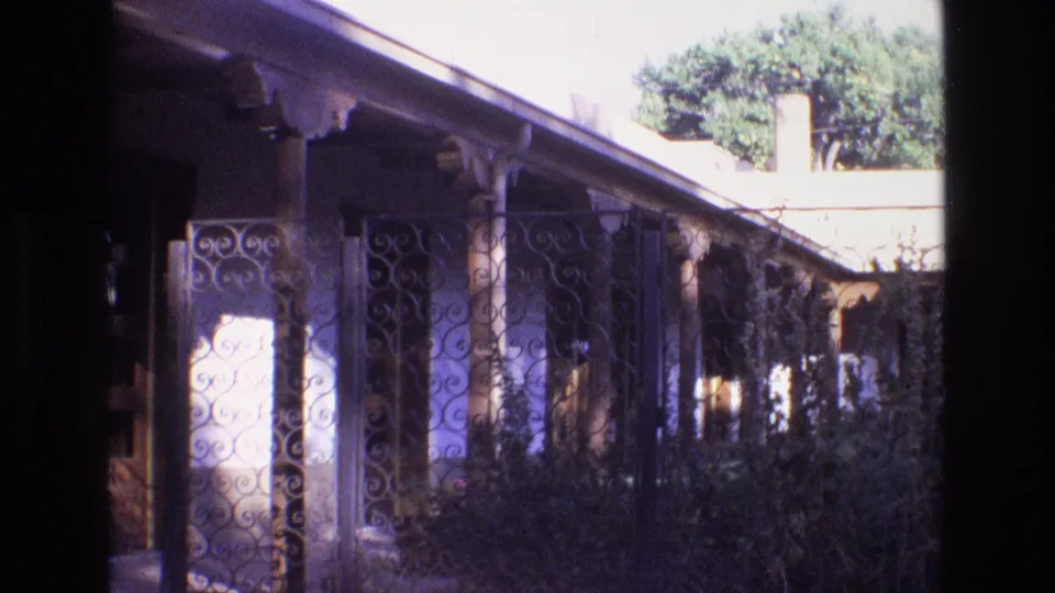 1973 New Mexico 8mm still showing a visitor's view down a shaded resort walkway with iron gate details, columns, greenery, and sunlit courtyard surfaces.