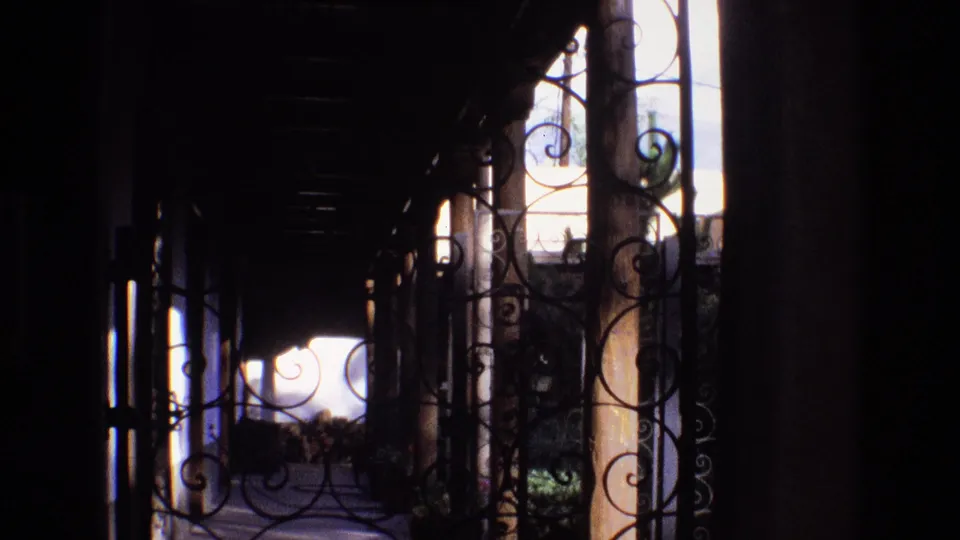 Final 1973 New Mexico frame looking through dark wrought iron scrollwork in a shaded resort corridor toward a bright courtyard opening.
