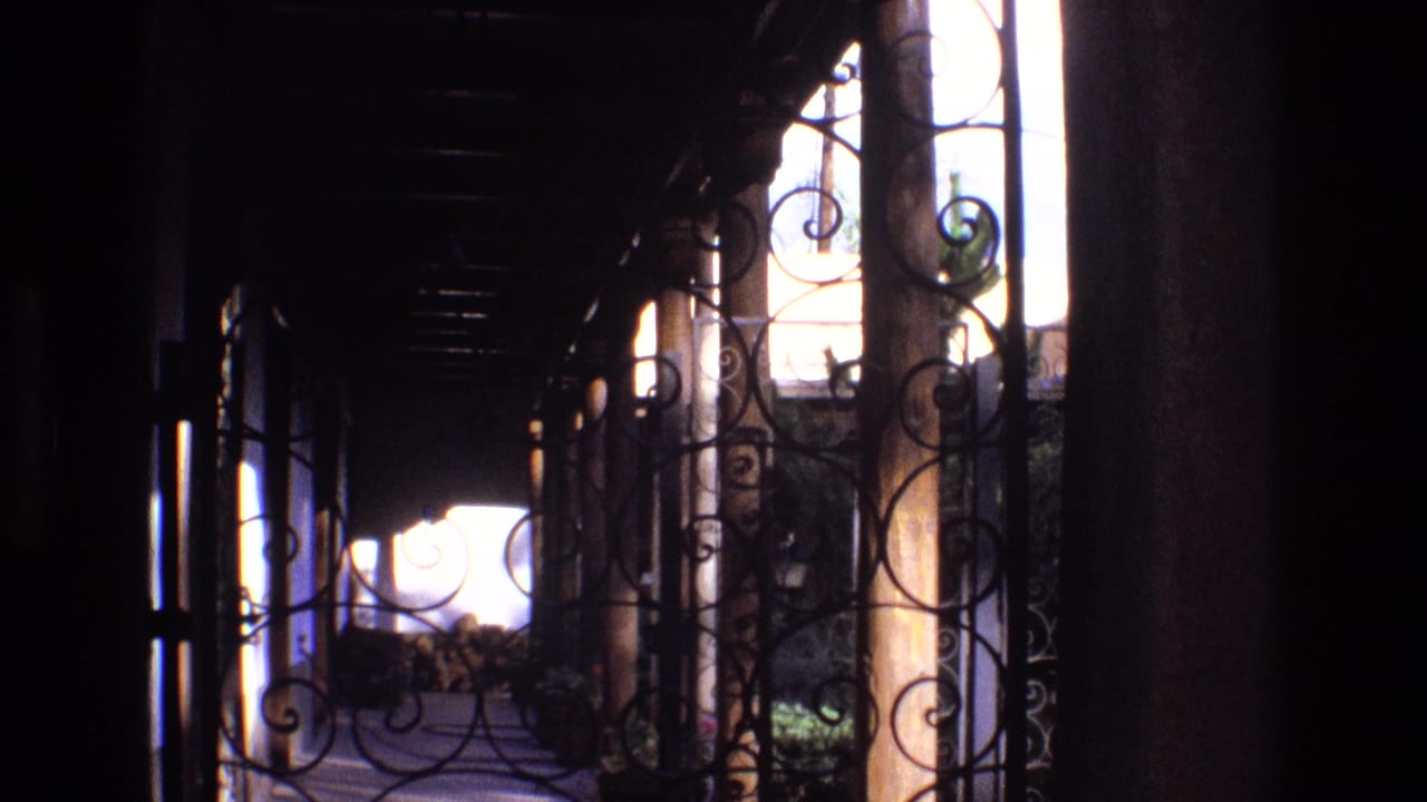 1973 New Mexico home-movie frame looking along a shaded porch through wrought iron bars and circular scroll designs toward sunlit outdoors.