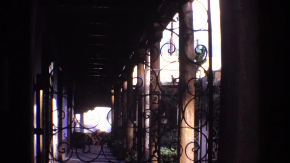 8mm frame from a 1973 New Mexico home movie showing a shaded residential porch, ornate iron railing, and high-contrast daylight beyond.