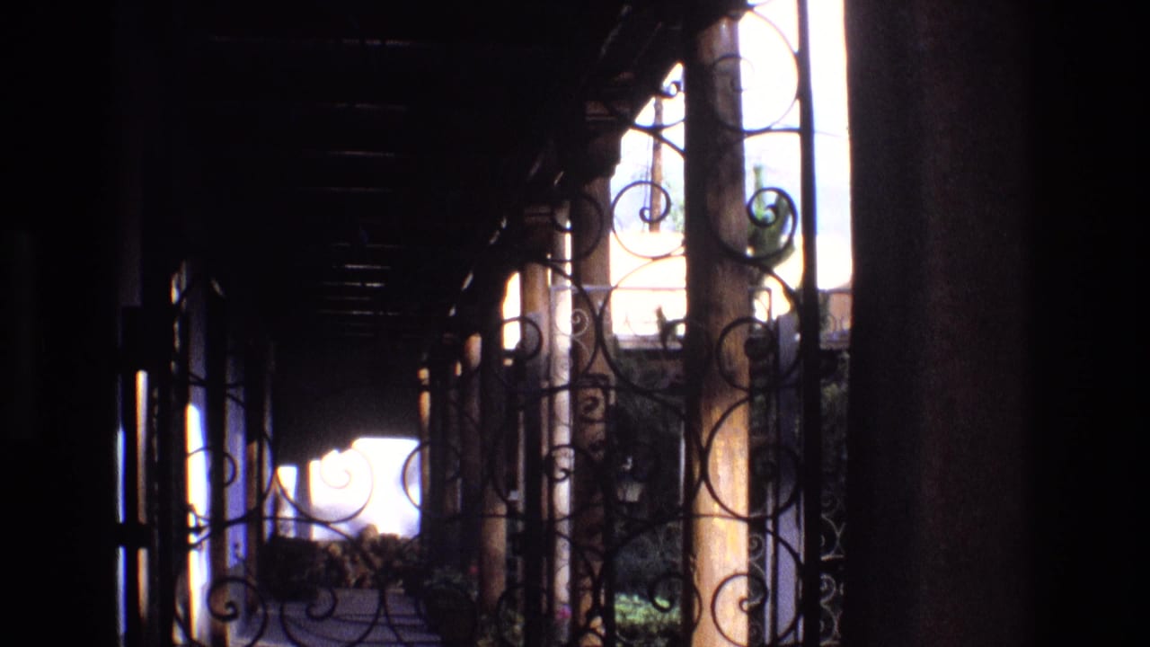 1973 New Mexico 8mm frame looking from a dark interior passage through decorative wrought iron and columns toward sunlit grounds.