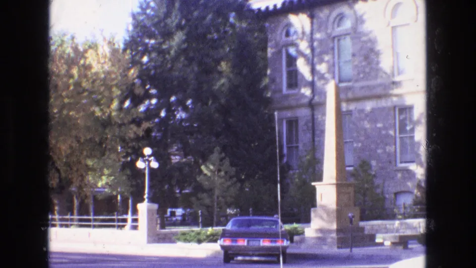 1973 New Mexico 8mm frame showing a formal yard with trees, a stone building facade, a tall obelisk-like monument, a lamp post, and a parked sedan.