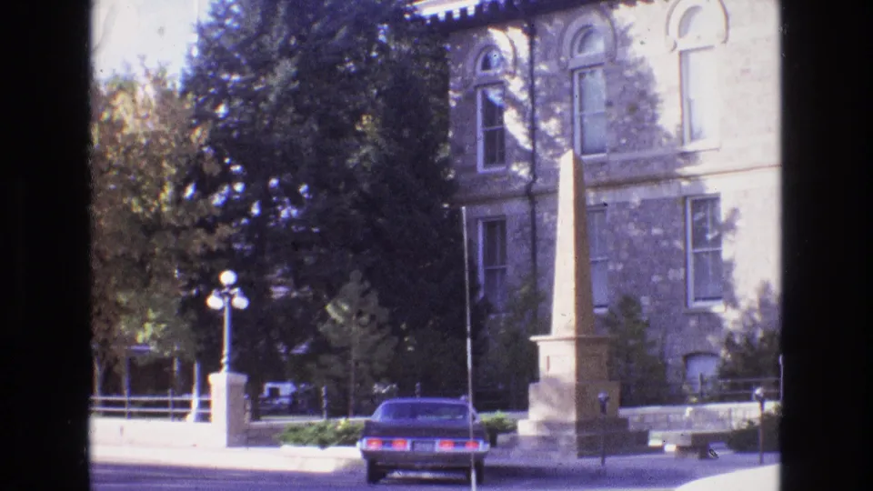 1973 New Mexico 8mm frame showing a stone mansion-style exterior with arched windows, an obelisk monument in front, a blue sedan at the curb, and trees shading the street.