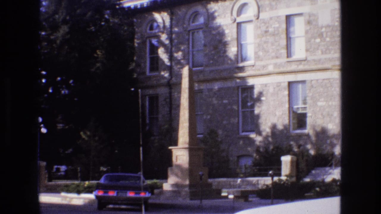 Obelisk monument and parked sedan in front of a stone building with arched windows and trees.