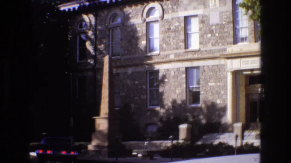 1973 New Mexico 8mm frame as the pan moves closer across the stone exterior, showing the obelisk, parked sedan, rectangular windows, entrance area, and low planting beds.