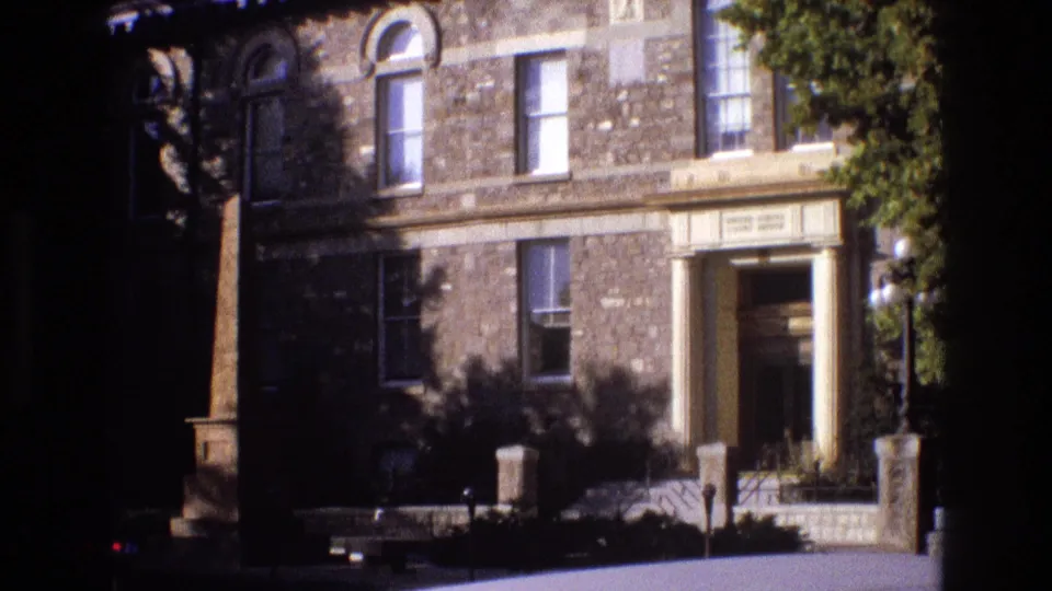 1973 New Mexico 8mm frame showing the right side of the stone building, including the recessed entrance, vertical columns, nearby obelisk base, tree canopy, and landscaped sidewalk.