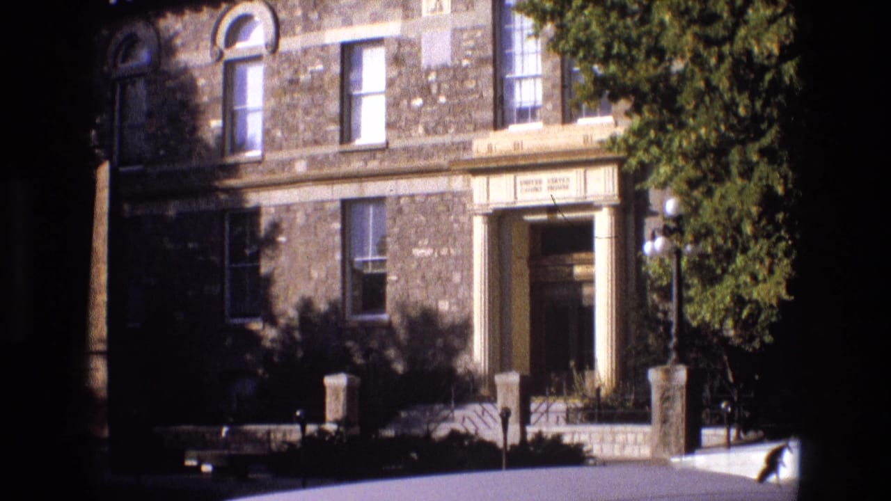 1973 New Mexico 8mm frame of a stone civic or institutional-looking exterior with a recessed entrance, tall windows, low posts, globe lamps, and leafy tree framing the right side.