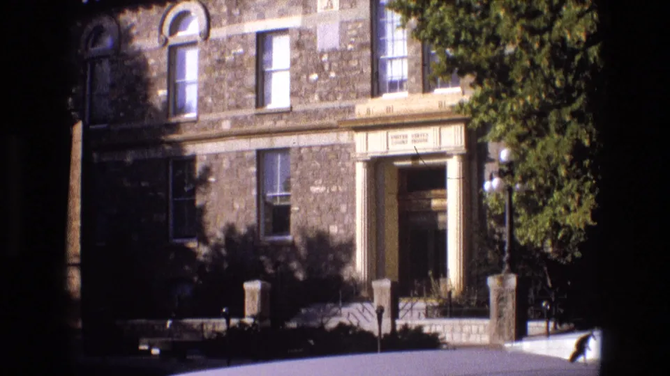 1973 New Mexico 8mm frame of a stone civic or institutional-looking exterior with a recessed entrance, tall windows, low posts, globe lamps, and leafy tree framing the right side.