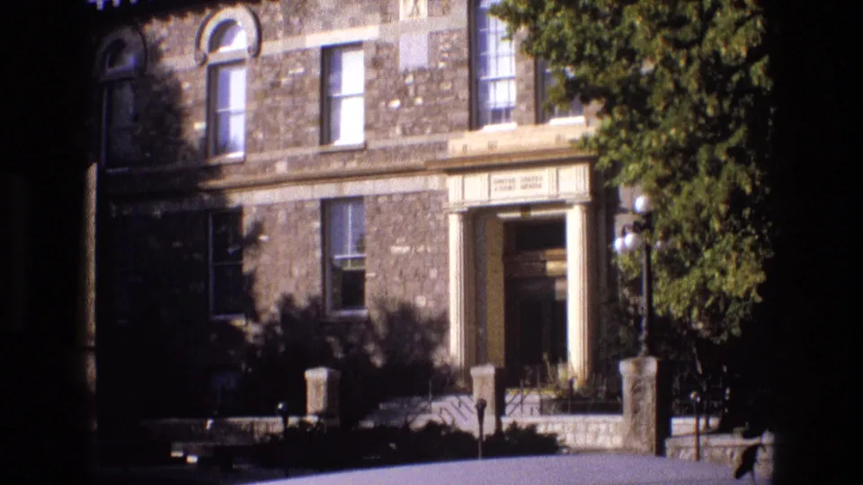 1973 New Mexico 8mm frame holding on the stone building entrance, with classical columns, arched window details, rectangular windows, globe lamps, masonry posts, and strong sunlight.