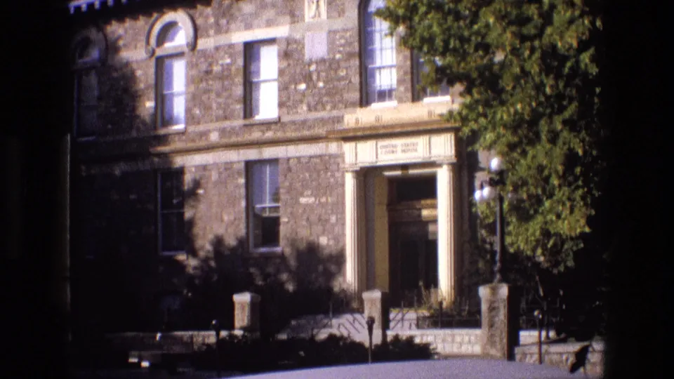 1973 New Mexico 8mm frame showing the same stone building exterior, including arched upper windows, a recessed columned doorway, globe lamps, low posts, and dense green foliage at right.