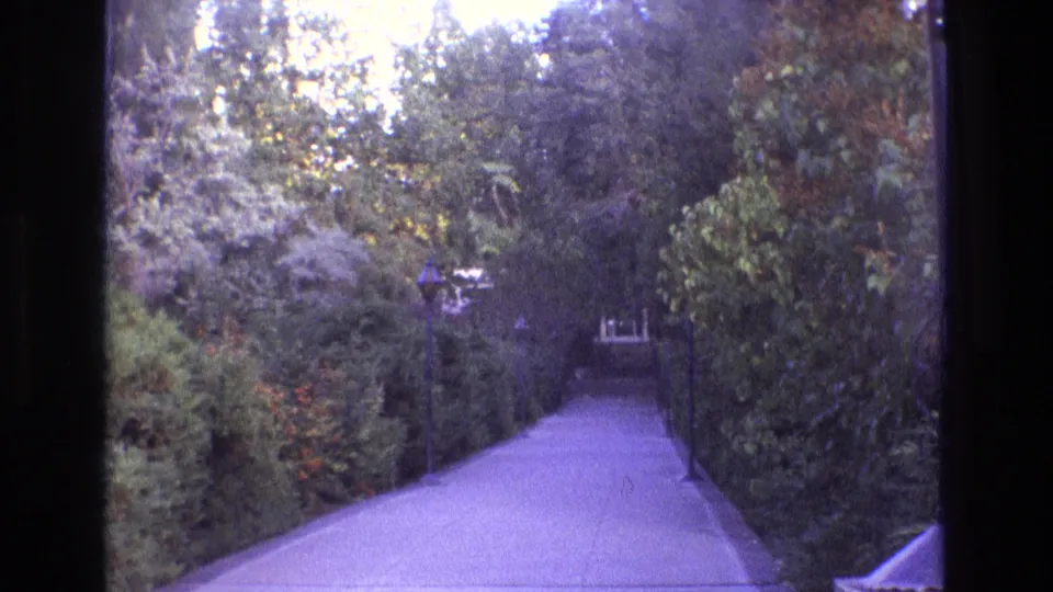 1973 New Mexico 8mm frame showing the garden path from a similar angle, with hedges, trees, and layered foliage enclosing the view.