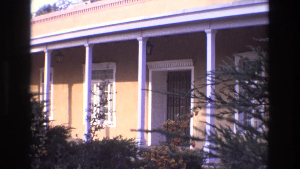 1973 New Mexico 8mm frame showing a yellow-toned house exterior with a covered porch, white columns, barred window, shrubs, and nearby trees.
