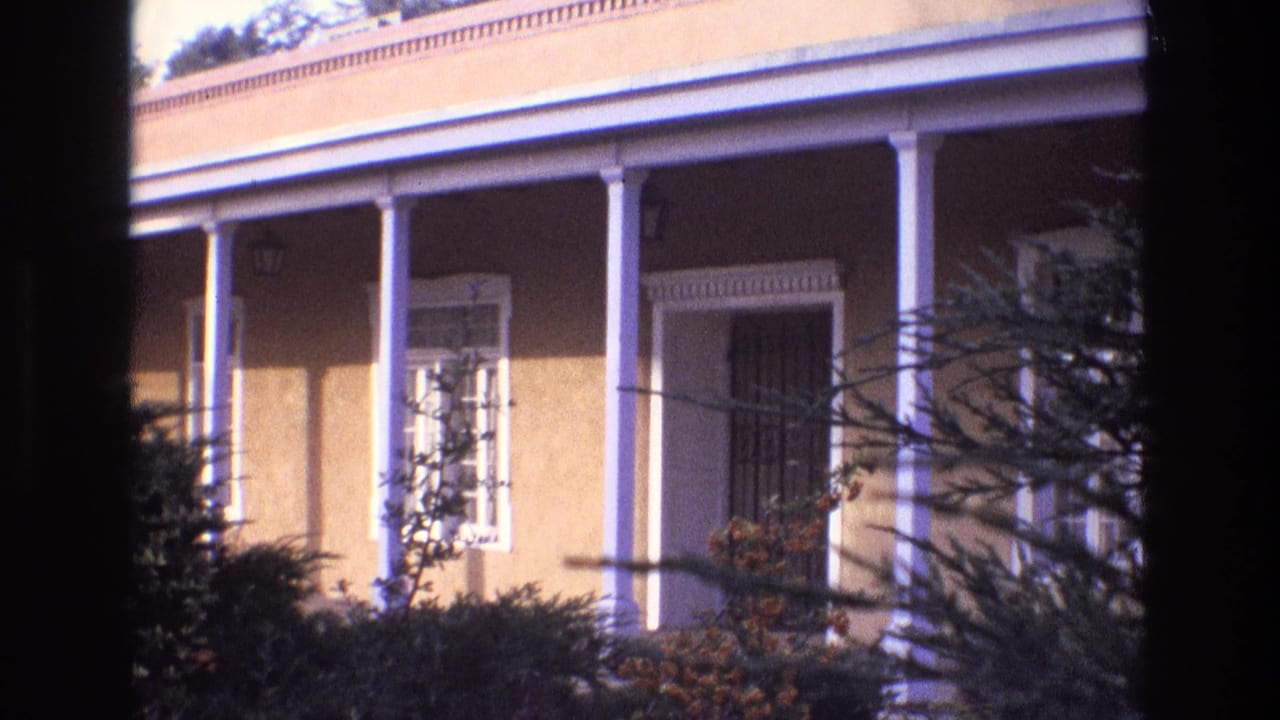 1973 New Mexico 8mm frame showing a sunlit stucco house exterior with white columns, a covered porch, window trim, and dark branches framing the view.
