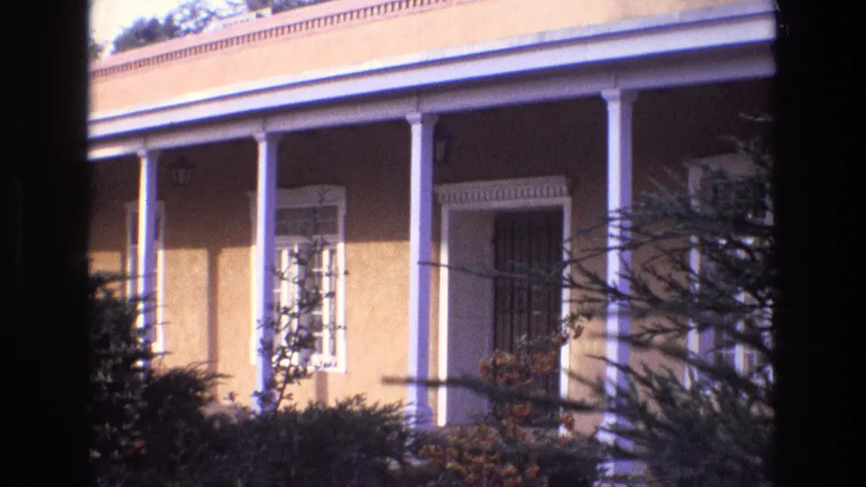 1973 New Mexico 8mm frame showing a sunlit stucco house exterior with white columns, a covered porch, window trim, and dark branches framing the view.