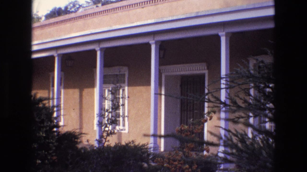 1973 New Mexico home-movie frame of a stucco house entry with white porch columns, a shaded doorway or grille, shrubs, and dark foliage partially framing the shot.