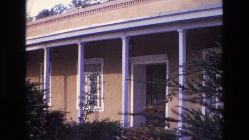 1973 New Mexico 8mm frame showing a steady exterior view of a stucco porch, white columns, a trimmed window or doorway, and foliage creating a dark natural vignette.