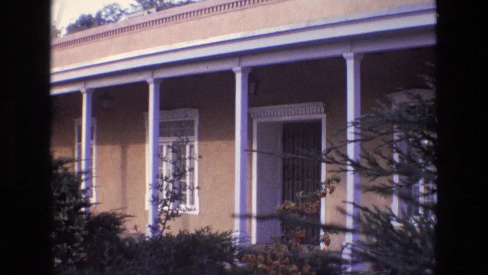 1973 New Mexico home-movie frame of a sunlit stucco house facade with white porch columns, a barred entry or window, shrubs, and branches crossing the foreground.