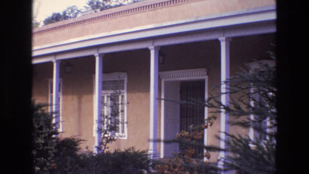New Mexico 1973 home-movie frame showing an adobe-style building with white porch columns, barred windows, foreground foliage, and part of the street.
